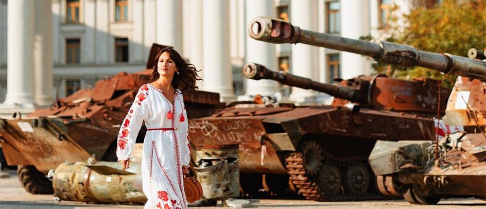 Woman in red and white dress walking by burned-out tanks in front of a large building with decorated columns